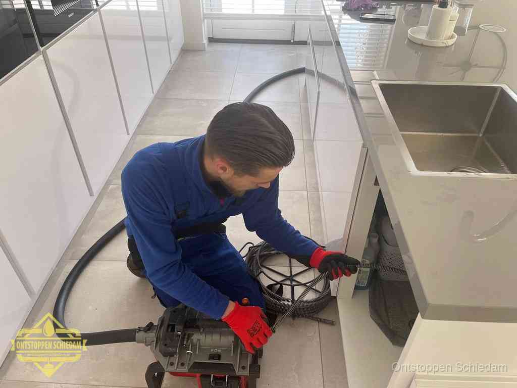 Een monteur ontstopt de keukenafvoer onder de spoelbak met een elektrische veermachine in Schiedam