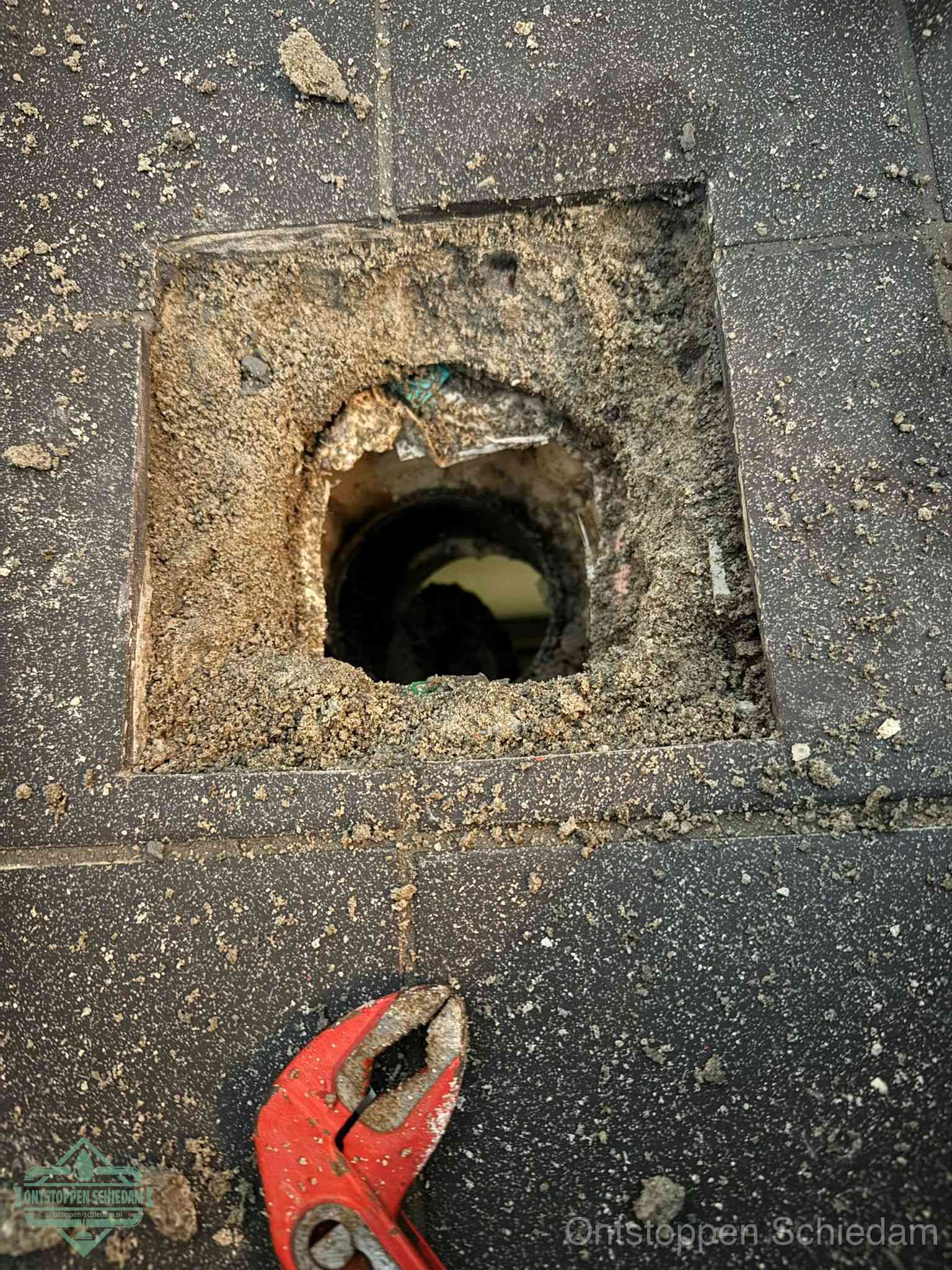 Een geopend rioolputje in een betontegelvloer met losgemaakt rooster en een zichtbare ronde afvoerbuis, het achterhalen waar de afvoerverstopping zit in Schiedam