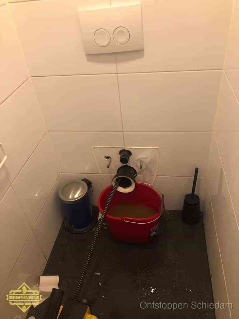 Een ontstopte wand-wc met verwijderde toiletpot waar een ontstoppingsveer in de wanduitgang verdwijnt, het achterhalen waar de verstopping zit in Schiedam
