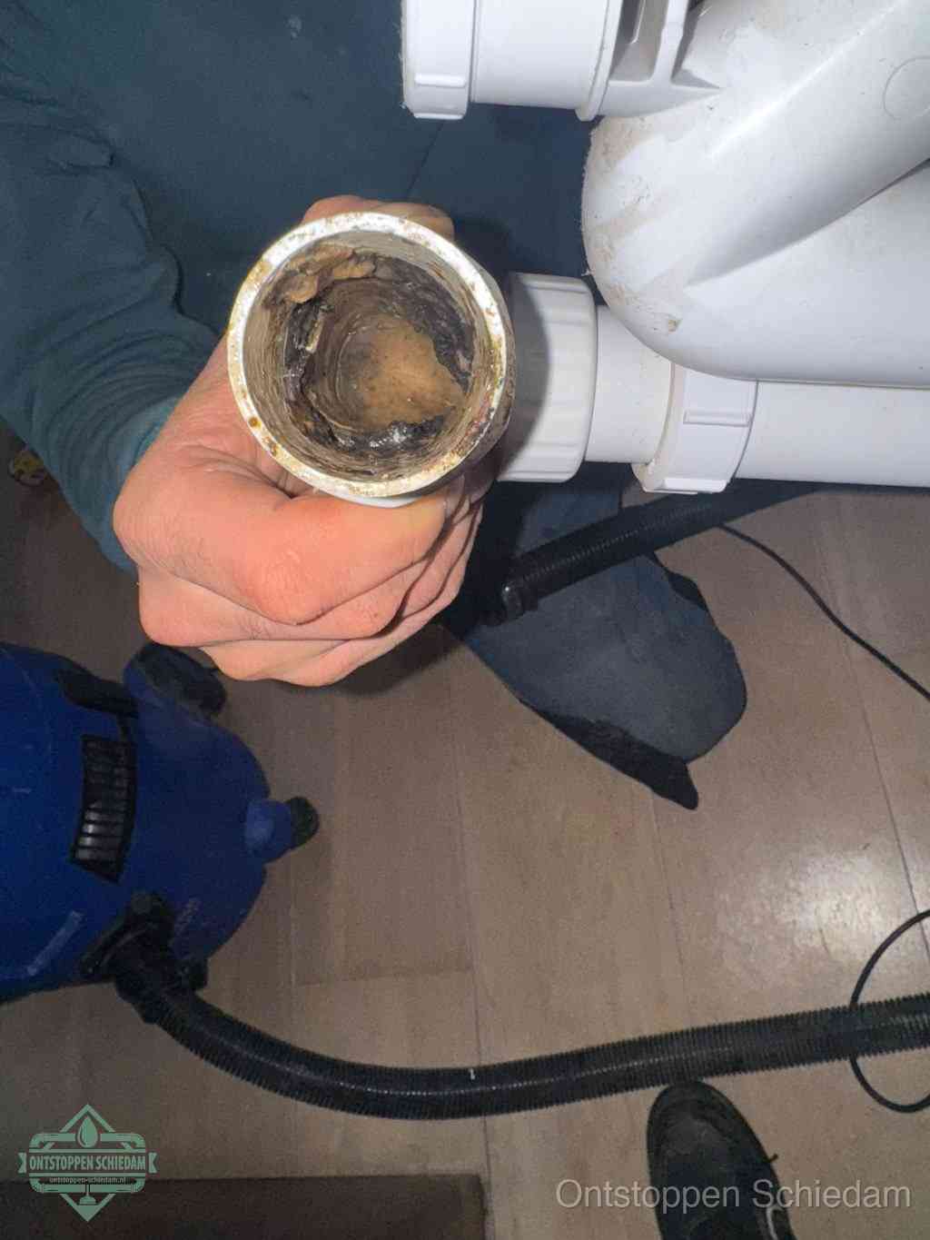 Een hand houdt een losgekoppeld PVC-afvoerbuisstuk vast dat van binnen volledig is dichtgeslibd met vet, vuil en aanslag, de meest voorkomende oorzaak van een verstopte afvoer in Schiedam