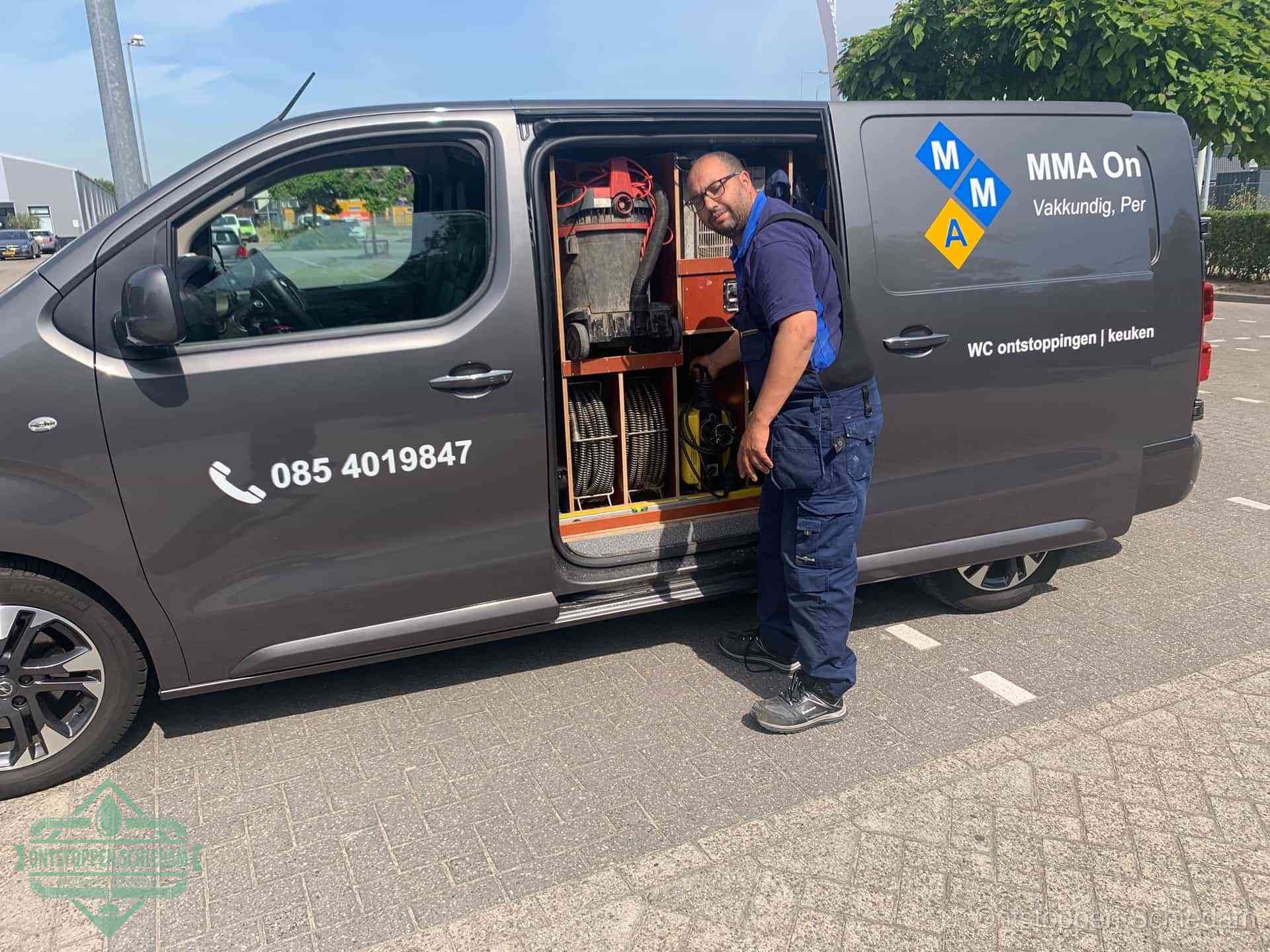 Een monteur bij de open schuifdeur van zijn bedrijfsbus op een parkeerplaats, klaar om de oorzaak van terugkerende afvoerverstoppingen aan te pakken in Schiedam