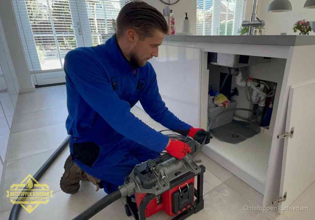 Een monteur voert een professionele elektrische ontstoppingsmachine in de afvoer onder de spoelbak tijdens het gootsteen ontstoppen in Schiedam